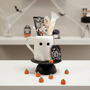 White ghost mug with black face and 'BOO' sign on a black stand, filled with treats and surrounded by Halloween-themed decorations.