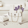 White mini purse tote with lavender-themed items including a bath salts, soap, lotion and spa gloves on a light surface
