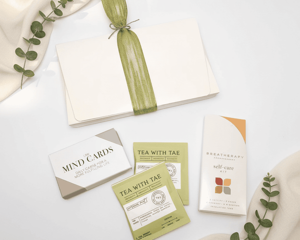 Collection of self-care items including cards, tea packages, and a box on a white surface with greenery
