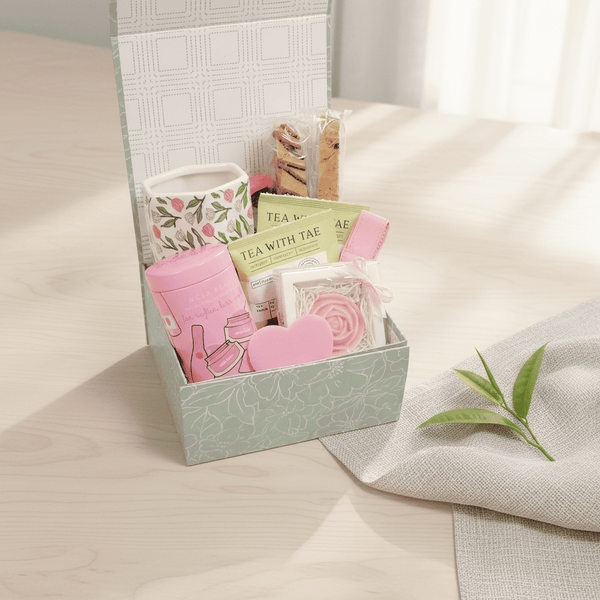 A floral mug, tea bags, a decorative rose soap, a lip care kit and a pink nail file in a decorative box on a light surface with a plant leaf