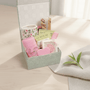A floral mug, tea bags, a decorative rose soap, a lip care kit and a pink nail file in a decorative box on a light surface with a plant leaf