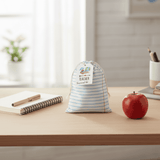 Striped drawstring bag labeled 'Teacher Survival Kit' on a desk with an apple and stationery items.