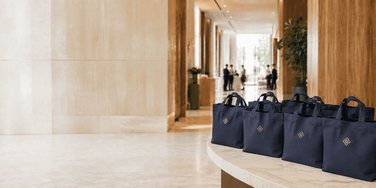 A row of navy blue canvas tote bags with gold logos lined up on a curved beige stone counter in a modern, upscale hotel lobby.