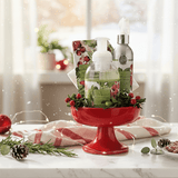 Celebratopia hostess set in Cranberry Wreath fragrance, with hand cream, room and linen spray, foaming hand soap and matching cocktail napkins in red pedestal bowl on a white marble countertop with Christmas decorations.