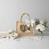Jute Photo Tote with a couple's image, adorned with a white bow, beside a vase of white roses, on a styled tabletop.