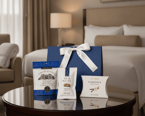 A gift set of gourmet snacks and treats arranged on a round dark wood table in a hotel room. The set includes a large blue gift box with a white ribbon, a blue and white bag of shortbread cookies, a white bag of nuts, and a white box of caramels.