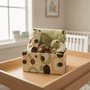 A baby gift set in a patterned storage box, featuring a green plush turtle toy, a folded green blanket, and a matching patterned cloth, displayed on a wooden changing table in a softly lit nursery.